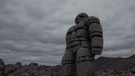 massive stone colossus rising from rugged terrain, constructed from weathered rock blocks, silhouetted against turbulent clouded sky, conveying primordial strength and enigmatic presence