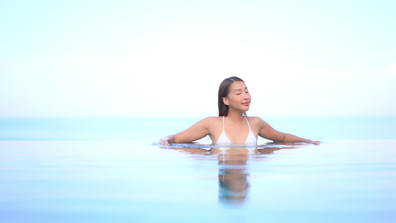 열대 스카이라인이 있는 인피니티 풀에 있는 매력적인 이국적인 여성이 수면, 풀 프레임과 합쳐집니다.