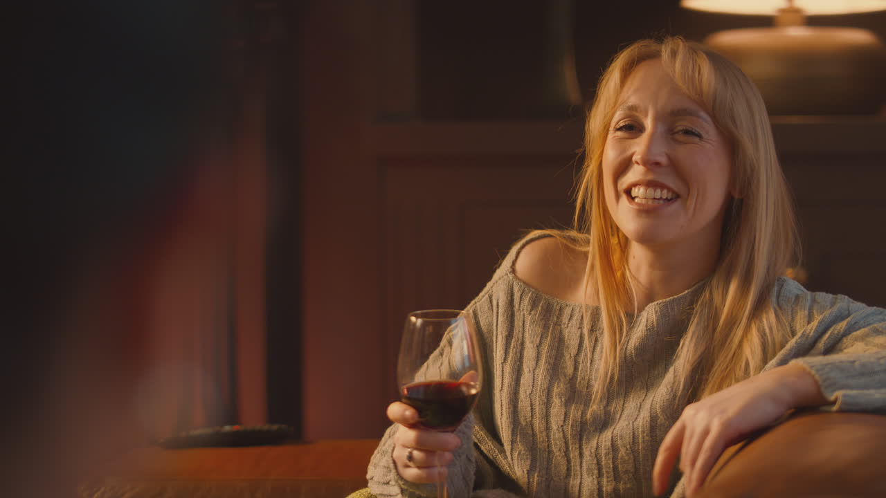 Two Female Friends Relaxing And Laughing On Sofa In Lounge With Cosy Fire Holding Glass Of Wine