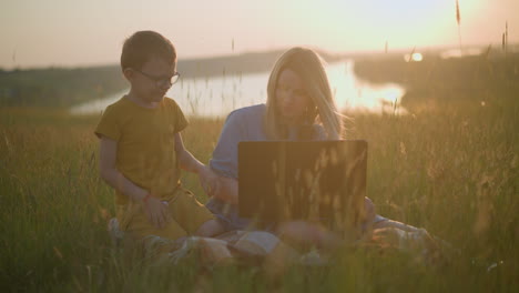 a woman in a blue gown sits on a scarf in a grassy field, working on her laptop during a peaceful sunset. a young boy dressed in yellow is seen walking towards her