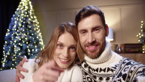 primer plano de una alegre pareja casada sonriente hablando en una videollamada en línea felicitar con las vacaciones en una habitación decorada con un árbol de navidad brillante en la víspera de año nuevo