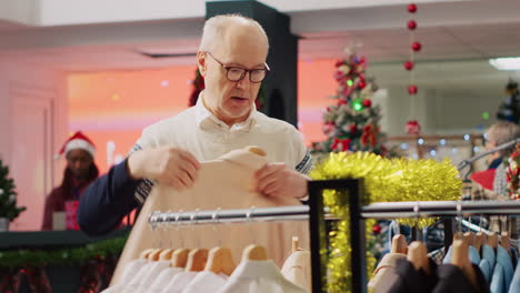 Stressed-older-man-browsing-through-clothes-in-festive-clothing-store-looking-for-family-gifts,-fed-up-with-Christmas-shopping.-Aged-client-picking-stylish-garment-for-son