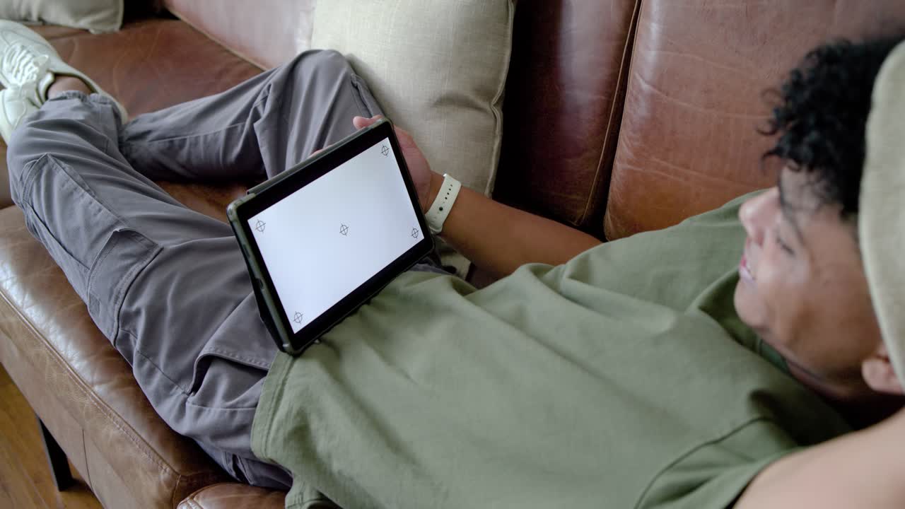Adolescente reclinado em um sofá de couro marrom, vendo a tela vazia de um tablet, alcançando e lendo