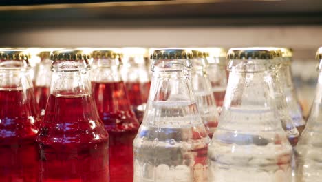 close-up of many glass bottles filled with red and clear liquids, possibly soda or other beverages, on a shelf in a refrigerator