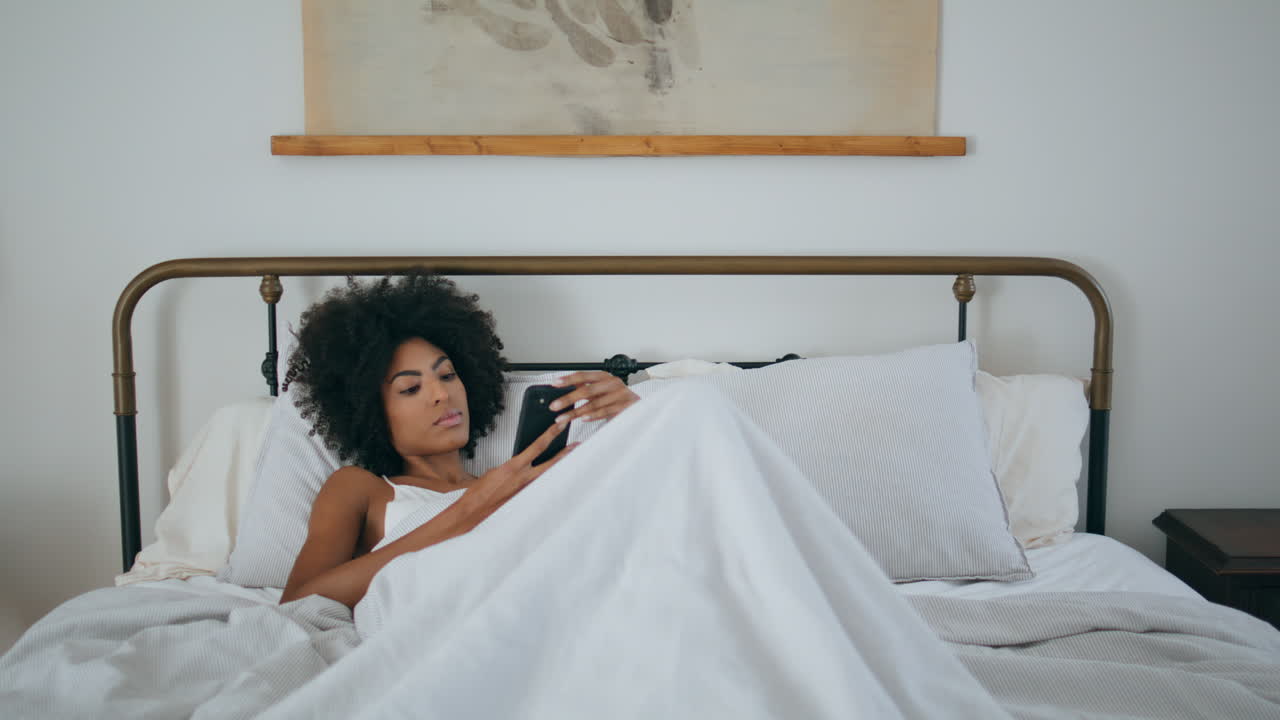 Modella delicata che legge lo smartphone in una camera da letto di lusso. Donna africana che tiene in mano il cellulare.