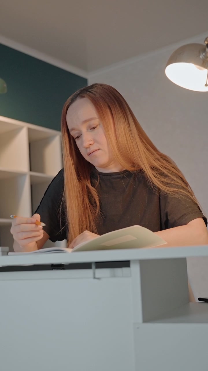 Girl records information into paper or digital medium. Subject briefly raises pen like object before resuming. Setting displays organized shelves and dual overhead lights