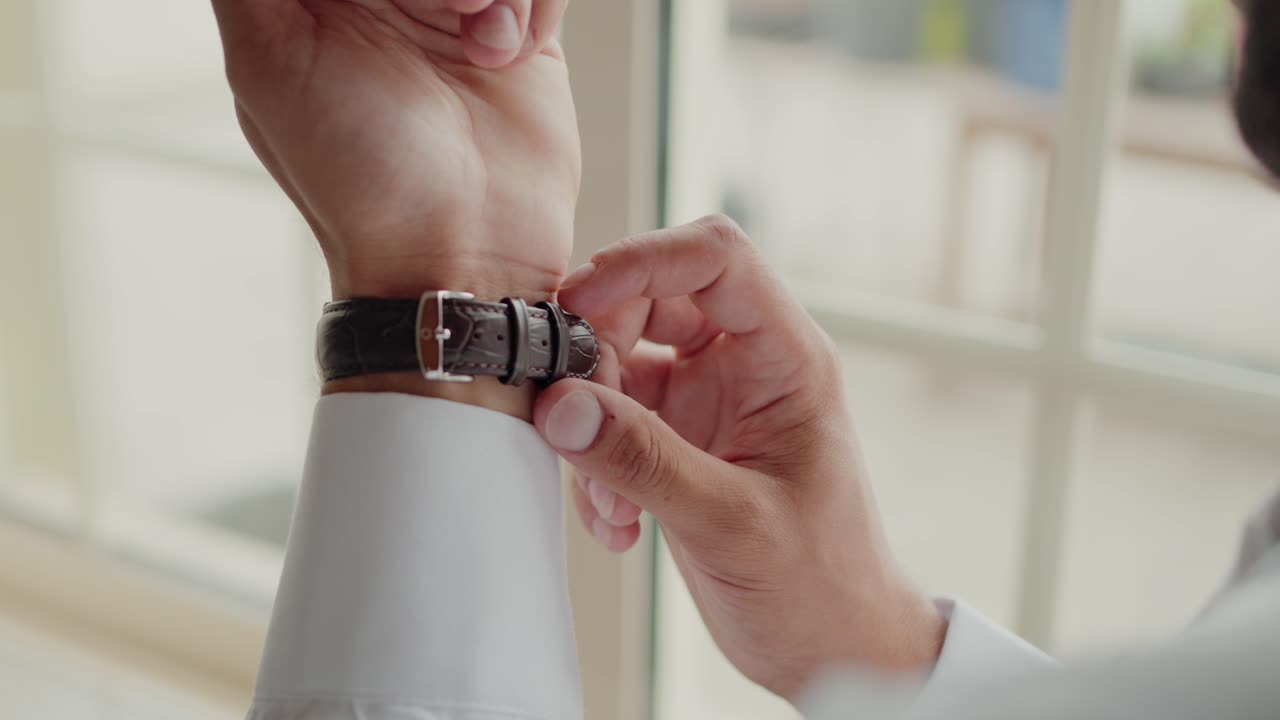 Man fastening a classic leather strap watch on his wrist near a window