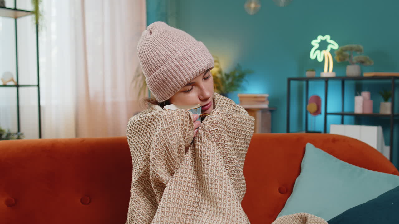 молодая нездоровая больная больная женщина, покрытая теплым одеялом, пьет кофе, замерзает от простуды и гриппа дома.