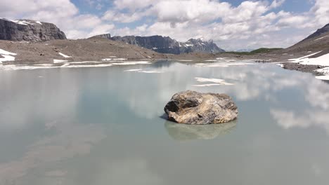 Solitary-rock-in-a-calm-alpine-lake-with-snowy-mountains-and-clear-skies-at-Klausenpass,-Switzerland