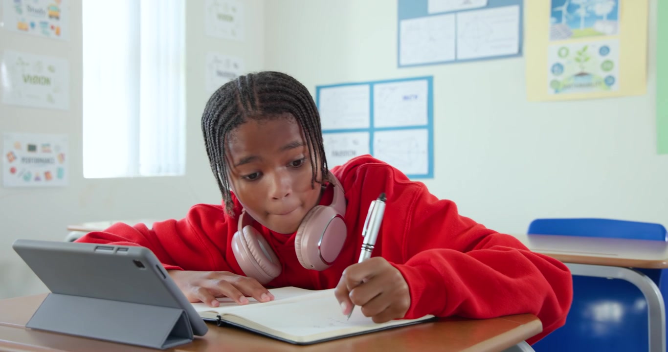 African Youth in red hoodie pink headphones glancing at tablet, writing notebook with pen in class