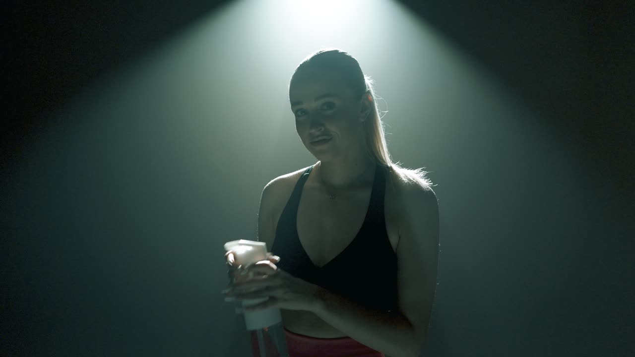 Blonde woman in pink shorts and black sports bra drinks water from transparent bottle, then smiles and looks at the camera. Upper body shot with dramatic rim light in a smoky gym environment