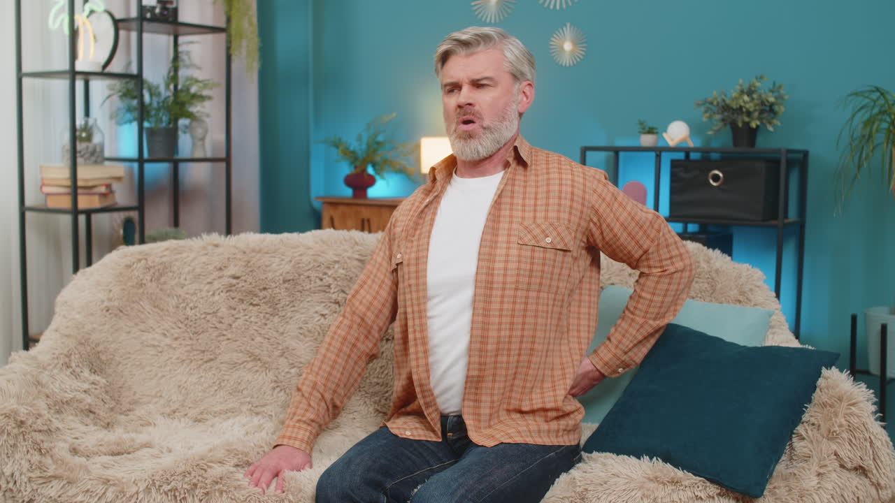 Mature man sitting on sofa holding lower back stretching and massaging back to relieve pain at home