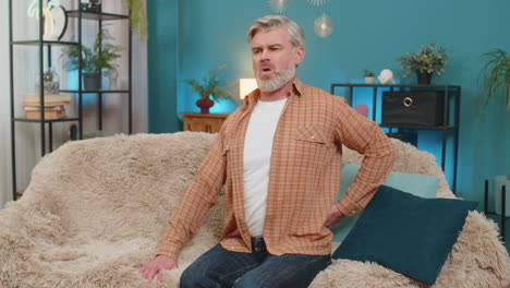mature man sitting on sofa holding lower back stretching and massaging back to relieve pain at home
