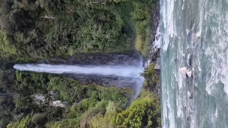 Vertical-De-Una-Cascada-Llamada-Thunder-Creek-Falls-Que-Desemboca-En-Un-Río-Rodeado-De-Una-Exuberante-Selva-Tropical-En-La-Costa-Oeste-De-Nueva-Zelanda