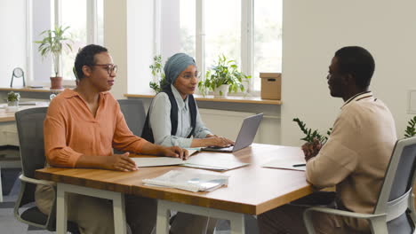 an woman and a muslim woman co workers interview a young man sitting at a table in the office 5