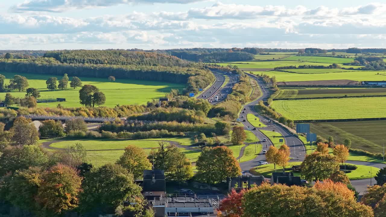 Drone panorama highlights A1(M) motorway and access roads crossing open countryside, farmlands, autumn foliage, woodlands and agricultural fields at Wetherby bypass, West Yorkshire, England