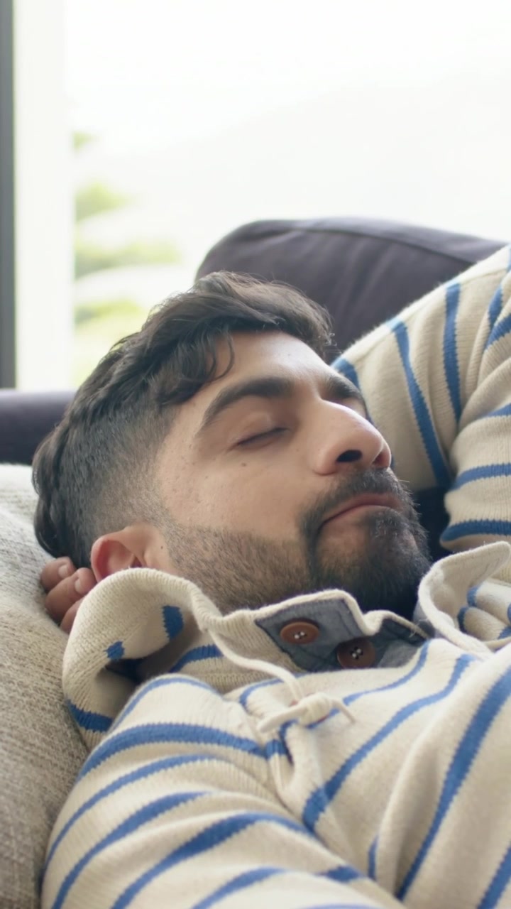 Vertikal video: Nærbillede af mand, der hviler på sofa ved vindue, iført pullover og nakkepude.