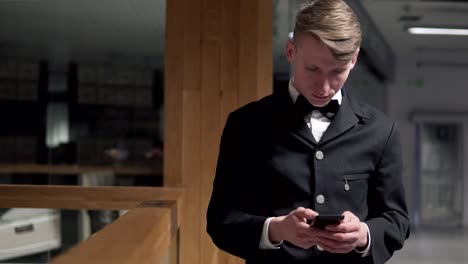 young attractive businessman standing near the stairs and uses a smartphone. he is dressed in and expensive suit with a bow tie