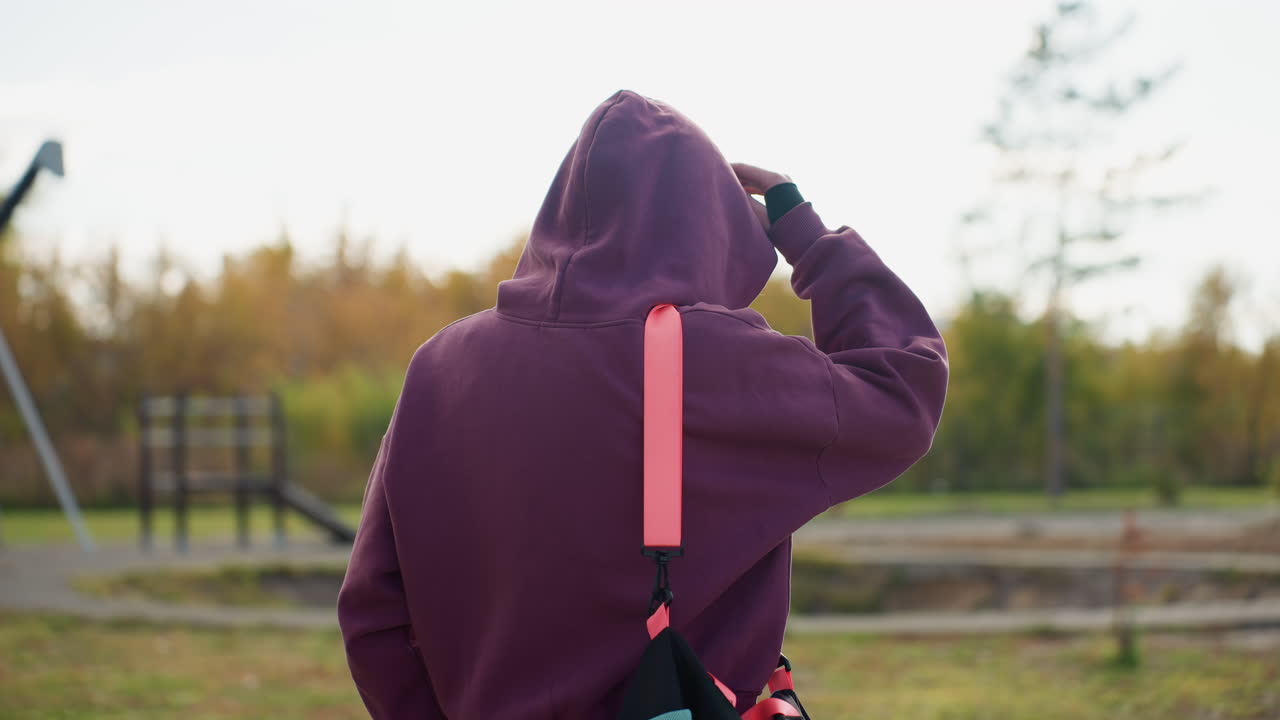 Back view of female figure carrying sport bag walking towards building through urban park pathway under bright daylight with autumn trees and modern facade backdrop capturing determined stride