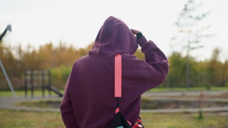 back view of female figure carrying sport bag walking towards building through urban park pathway under bright daylight with autumn trees and modern facade backdrop capturing determined stride