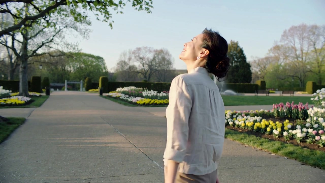 Walking woman in light shirt spotting fountain turning up pausing holding phone in public garden
