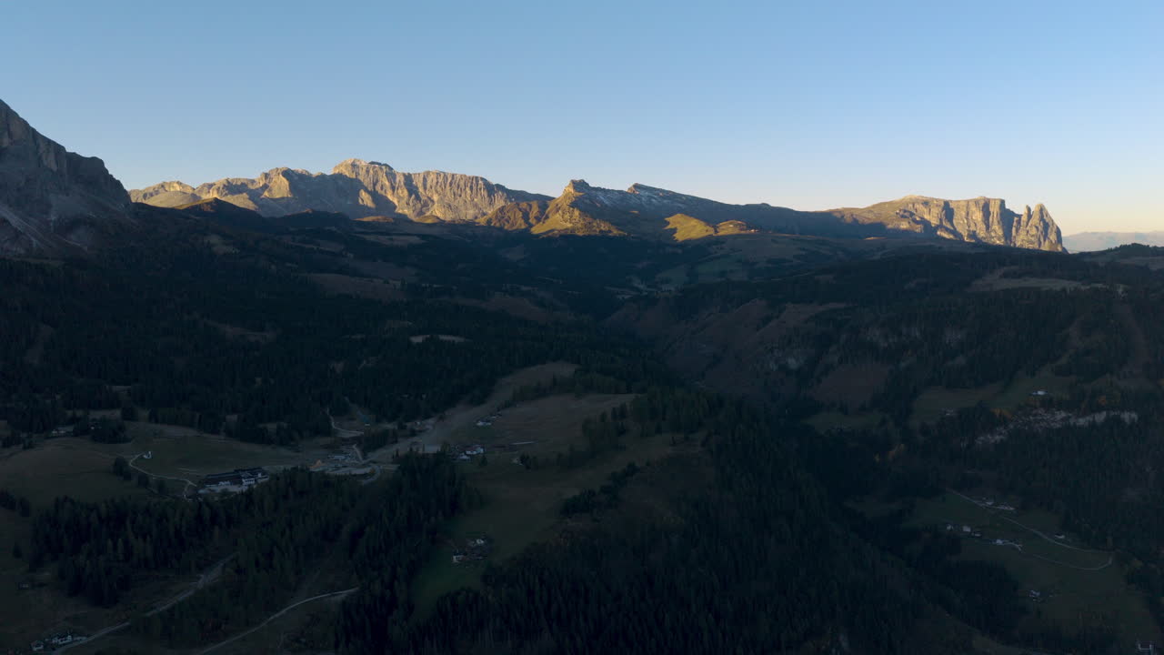 Pemandangan udara sinematik Dolomites Italia saat matahari terbit, puncak berbatu terjal di atas perbukitan alpine yang subur dan chalet yang tersebar, pemandangan alam lanskap perjalanan gunung yang tenang