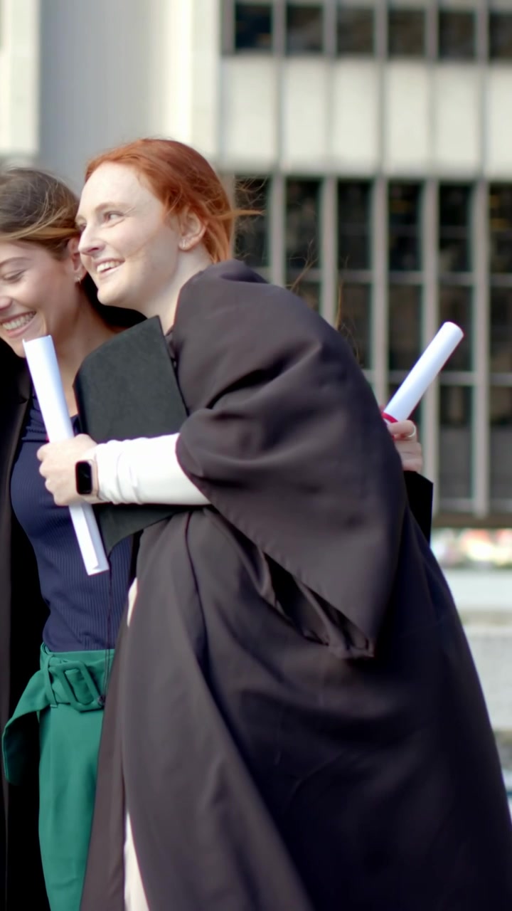 Video verticale: Laureata rossa in toga nera che cammina attraverso la piazza dopo una foto con tocco e diploma.