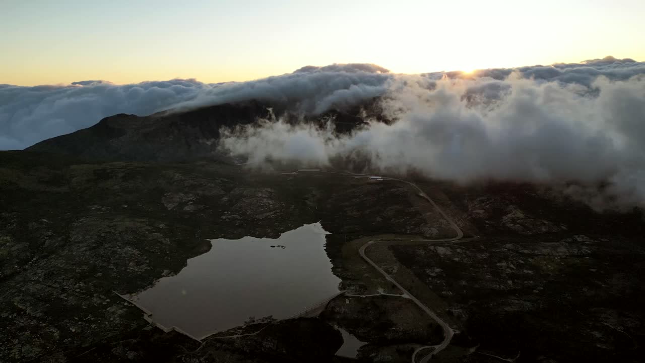 From an aerial vantage point, behold a panoramic view of Portugal's highest peak in the Serra de Estrella, where dense cloud formations share the sky with a backdrop of a rising sunset