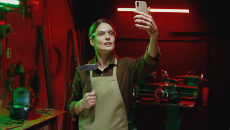 Caucasian-welder-woman-in-goggles-and-holding-hammer-making-a-video-call-on-smartphone-in-metal-workshop