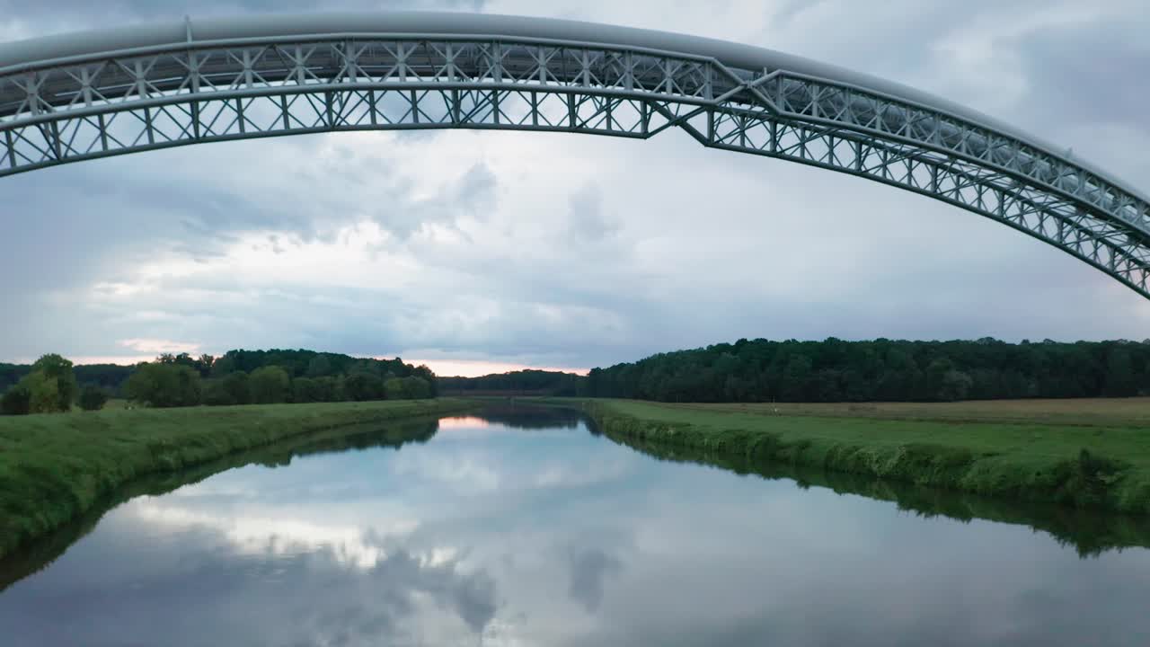 Flying backwards under two bridges for gas pipes running across the calm river Moravia. Aerial sunset footage of symmetrical gas pipe bridges and clouds reflecting in water