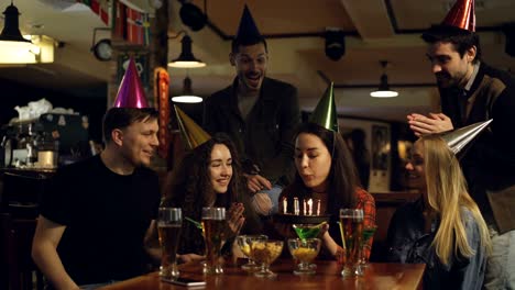 una joven morena está haciendo deseos y soplando velas en el pastel de cumpleaños mientras celebra el cumpleaños en un café con amigos. personas felices con sombreros de fiesta están aplaudiendo.