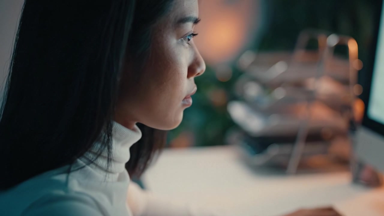 薄暗い環境でコンピューターに向かい、集中力と決意をもって仕事に取り組む若い女性。