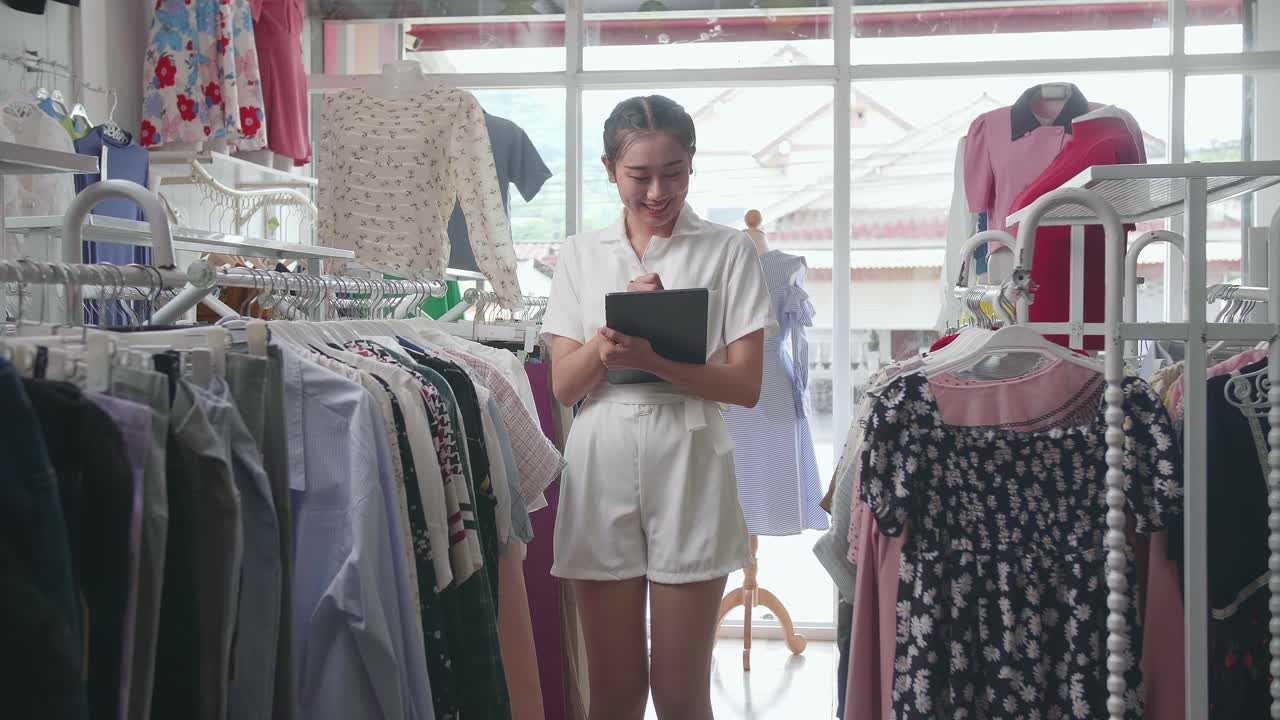 propietaria asiática de una tienda de ropa tomando notas en la tableta y sonriendo a la cámara mientras camina comprobando las existencias en los rieles