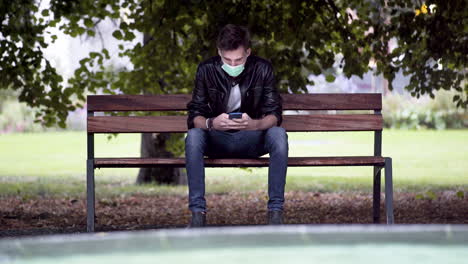 A-young-man-in-jeans,-white-T-shirt-and-a-black-jacket,-wearing-a-green-Covid-19-facemask,-sitting-on-a-park-bench,-waiting-for-someone,-taking-his-phone-and-unsuccessfully-trying-to-call-them,-4k