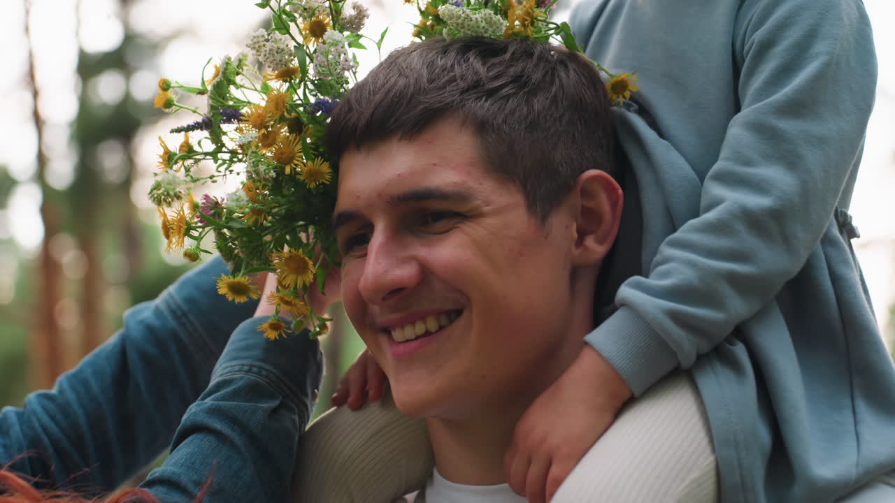 Young man carries little sister on shoulder in peaceful forest while smiling mother gently places bright flower bouquet on his shoulder, showing warmth, affection, and joyful family bond