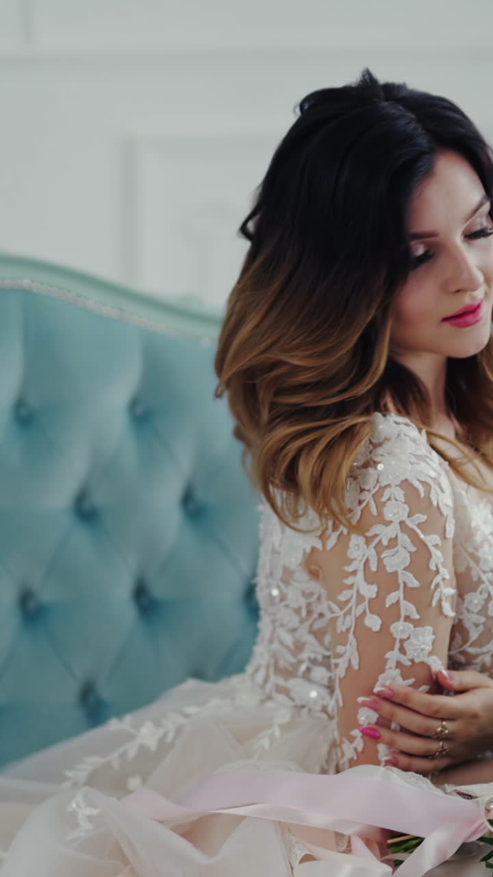 Lovely bride in a nice wedding dress with flowers is sitting on the luxury couch. Beautiful model girl with perfect makeup is posing to camera on a modern studio background. Vertical video