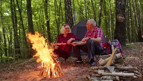 Happy-senior-man-and-woman-talking,-cooking-frying-sausages-over-campfire-in-evening-wood,-camping