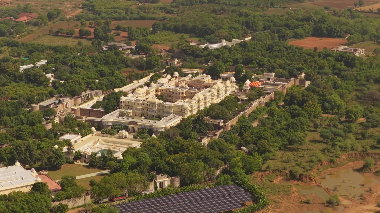 Una ripresa cinematografica dall'alto di un maestoso palazzo bianco in India, circondato da alberi verdi lussureggianti e moderni pannelli solari, che mostra un connubio tra patrimonio e sostenibilità