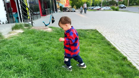 cute toddler boy in a motley shirt lets go mom's hand and steps on the grass. kid walks by the grass to shop windows.