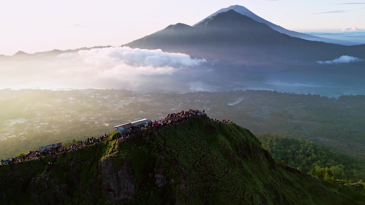 Nascer do sol sobre um cume de montanha lotado com caminhantes ao longo de uma trilha exposta no cume, cena contraluz com névoa brilhante revelando um lago de cratera e um pico vulcânico, Bali, Indonésia, tomada aérea de drone com aproximação