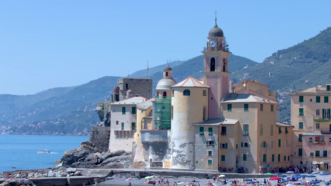 Inquadratura ampia statica di una città costiera mediterranea con edifici color pastello e spiaggia di ciottoli.