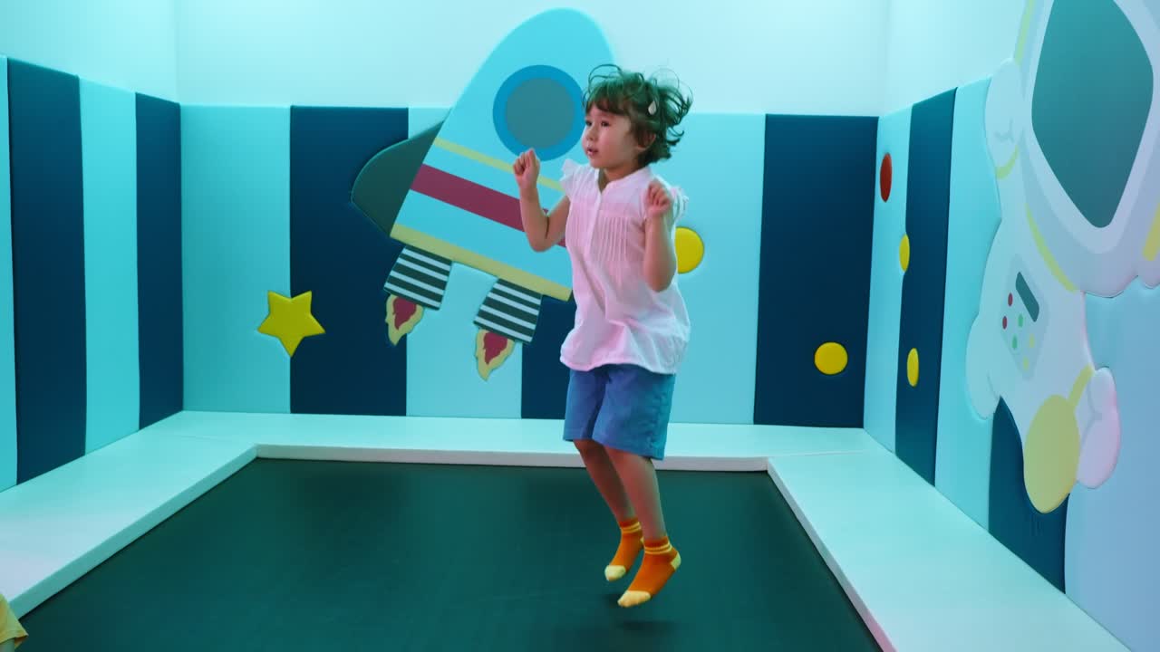 A 5-year-old girl smiles and jumps on a small indoor trampoline in a brightly lit room decorated with a space theme and a rocket on the wall