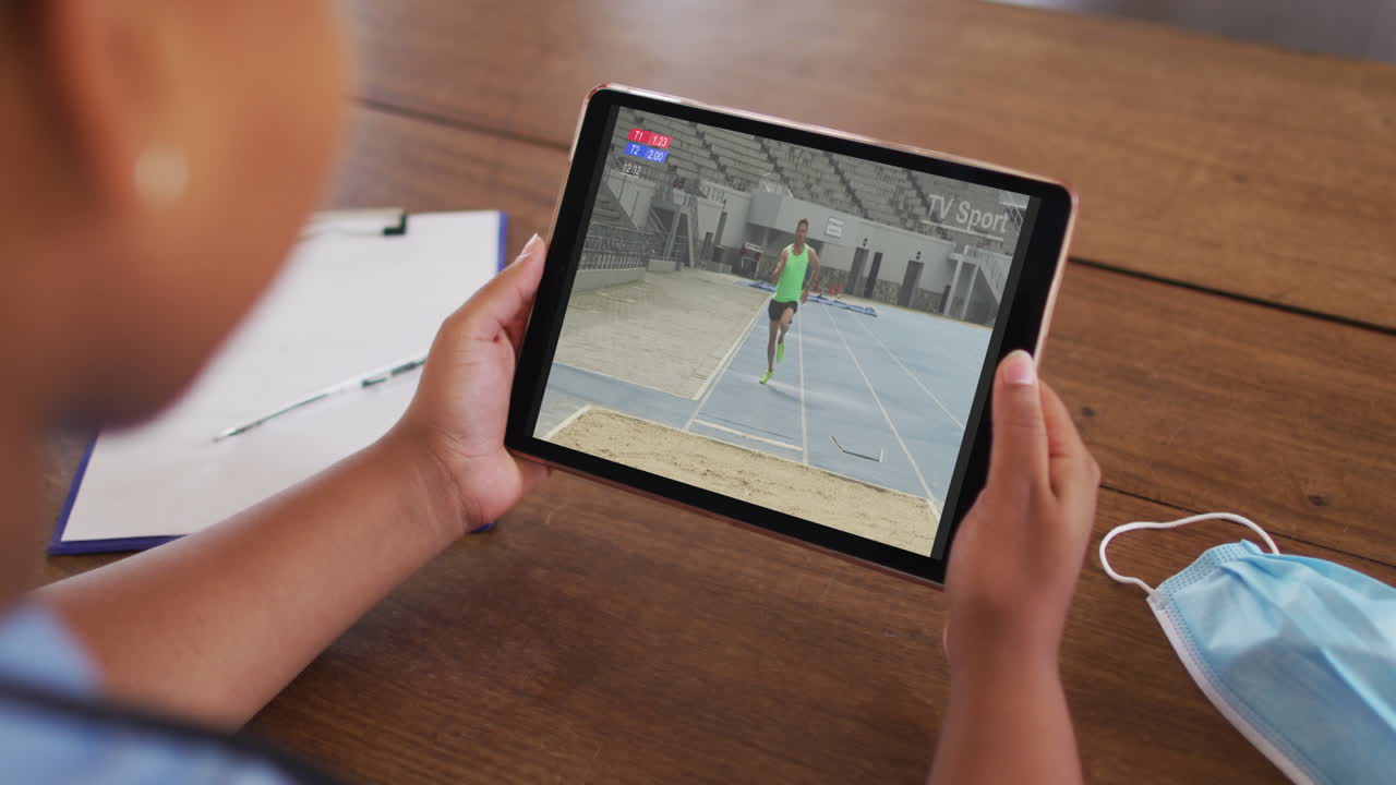 태블릿에서 육상 멀리 점프 이벤트를 시청하는 얼굴 마스크와 함께 테이블에 앉아있는 여성의 복합