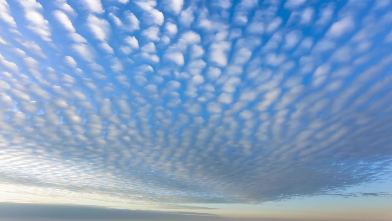 Dense rippled cloud layers sweep across bright blue sky above soft horizon glow, forming calm airy weather scene with natural daylight, textured depth, and broad copy space for seasonal, travel, and