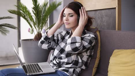 Gorgeous,-young-woman-relaxes-sitting-on-couch,-puts-on-headphones-and-turns-on-music-on-laptop.-Smiles-and-moves-to-the-rhythm-of-music.-Slow-motion