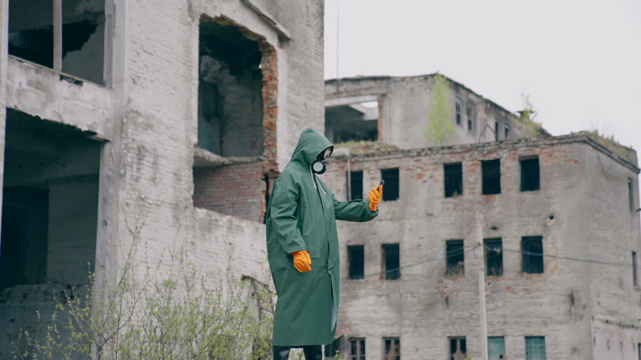 Human in protective suit and gas mask in abandoned place. Survivor holding a mobile phone while standing in ruined city. Post-apocalyptic world after a nuclear war.