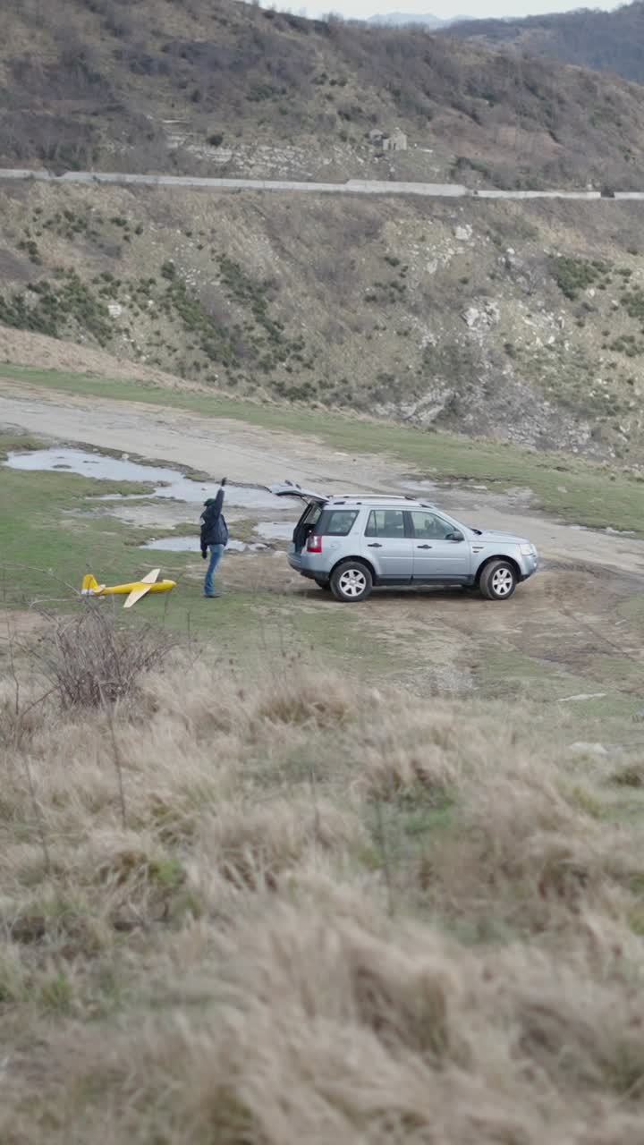 Man launching model airplane near car in mountainous landscape