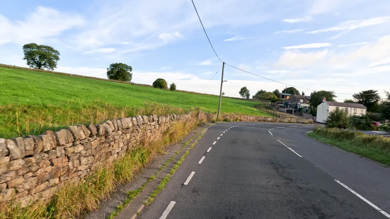 A vehicle travels along a rural English road bordered by stone walls and fields, entering a small village under soft evening sunlight with minimal traffic