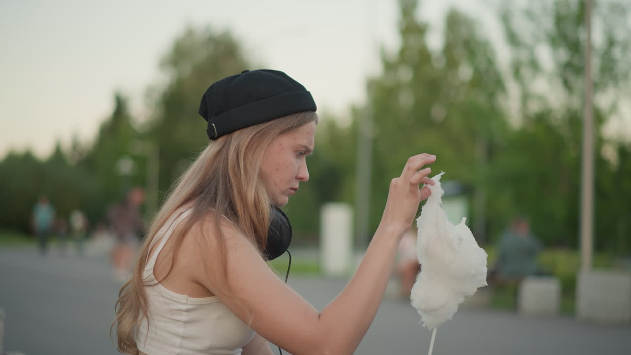 ragazza caucasica adolescente mangia zucchero filato all'aperto, cuffie e berretto visibili, atmosfera informale di fiera nel parco con venditori e passanti, mani che prendono la dolce ovatta, morsi giocosi e piccole fuoriuscite, spontanea