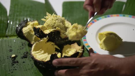 Roasted-breadfruit-in-open-fire,-man-spooning-out-the-cooked-roasted-breadfruit-on-banana-leaf-and-putting-in-plate,-traditional-Seychelles-food,-Mahe,-Seychelles-4k-24fps-009